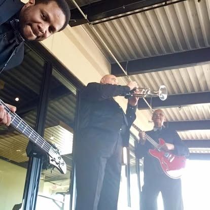 Jazz band performing outdoors: trumpet, bass guitar, electric guitar, and smiles.