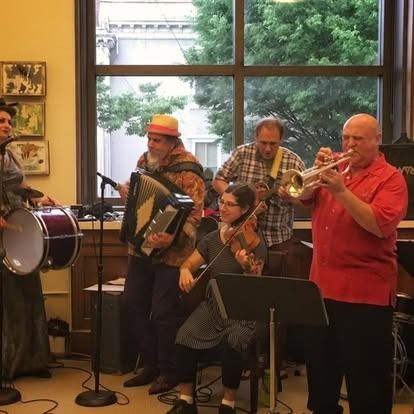 A band performs indoors: drummer, accordion, violin, guitar, trumpet. Bright lighting and large window.
