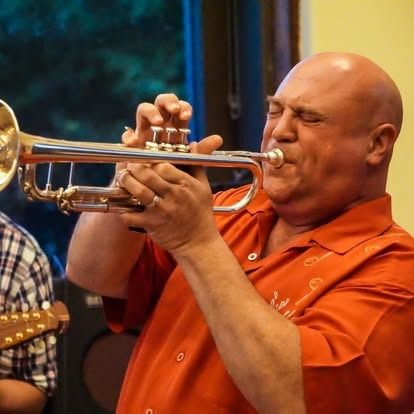 Bald man playing a silver trumpet, wearing an orange shirt, indoors.
