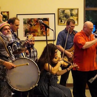 A band of seven musicians performing indoors. Instruments include drums, violin, accordion, and trumpet.