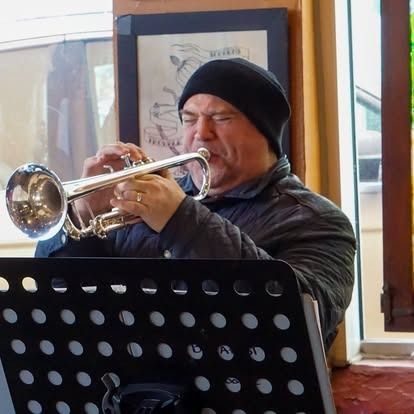 Man in black beanie plays a silver trumpet; music stand in front.