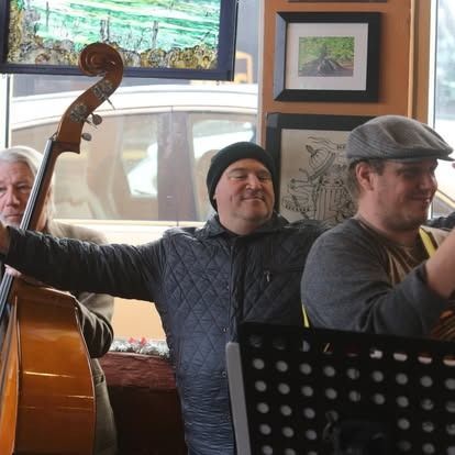 Three musicians perform indoors: bassist, banjo player, and a singer in a black beanie, smiling.