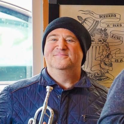 Man wearing a black beanie and quilted blue jacket, smiling, holding a trumpet.