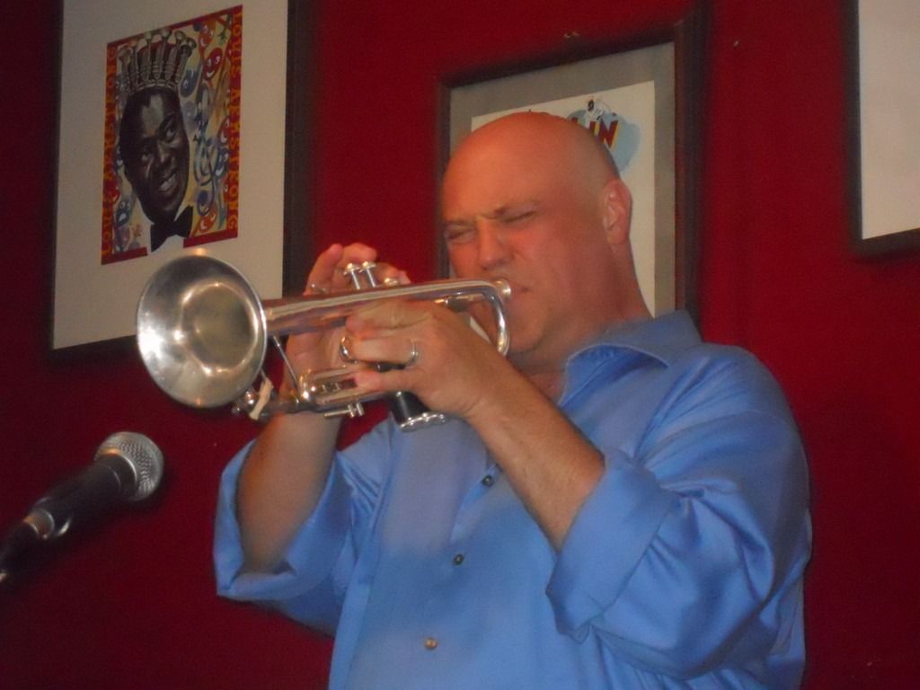 Bald man in blue shirt playing a silver trumpet, red-walled bar with jazz artwork.