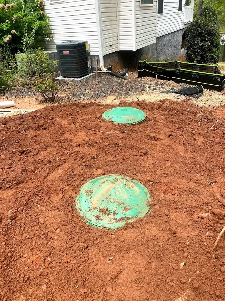 Two green septic tank covers in red dirt near a white house with a black AC unit.