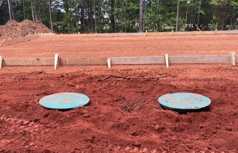 Two green circular covers in red dirt near a construction site foundation.