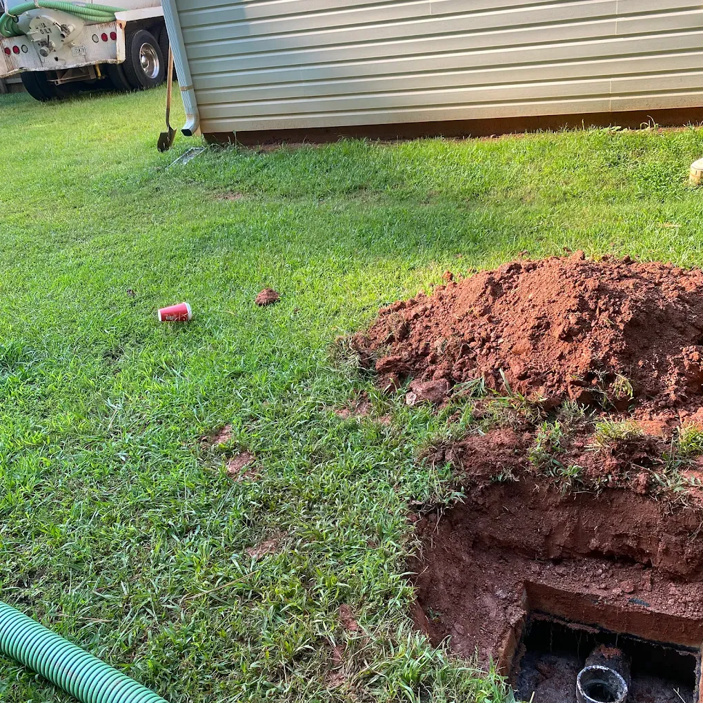 Hole dug in grass next to a house with a pile of dirt and a septic tank opening.