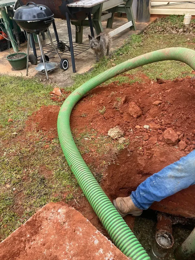 Green hose in a hole with a person's leg and a small charcoal grill in the background.