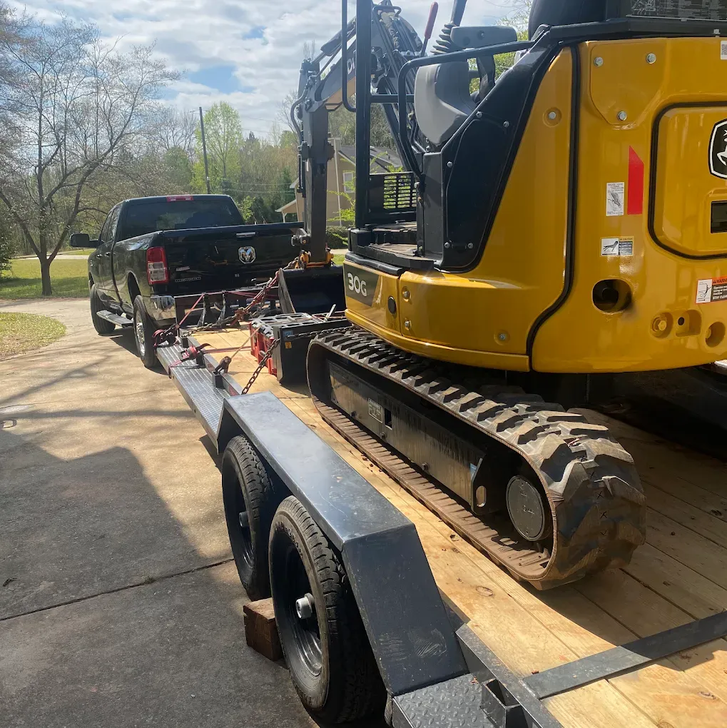 A black pickup truck towing a flatbed trailer with a yellow John Deere excavator.