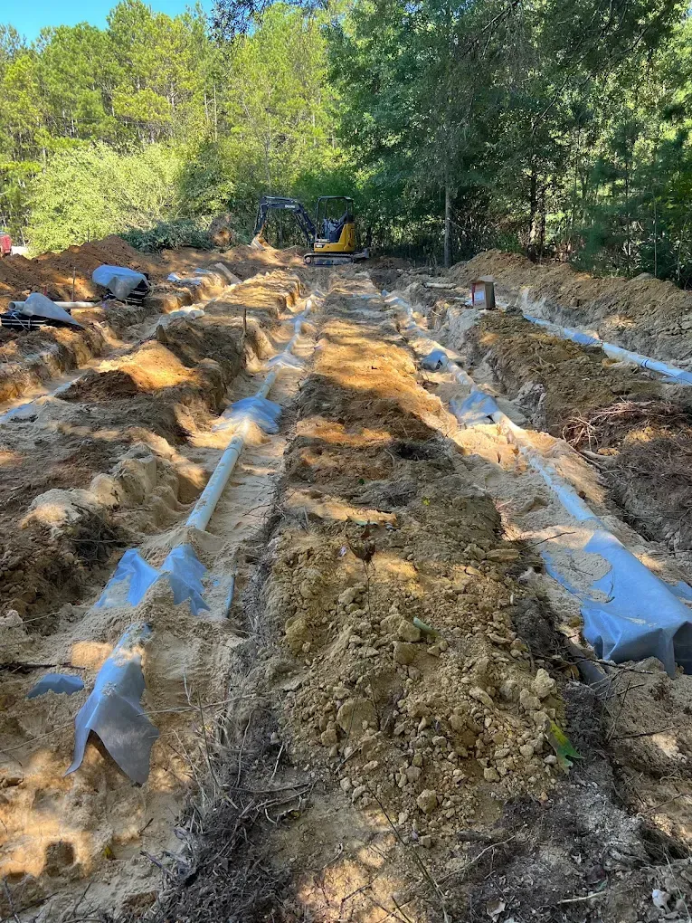 Trenches dug in soil, with gray pipes and a small excavator in a wooded area.