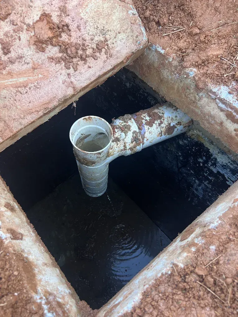 Open septic tank with pipe entering, dark liquid inside. Brown dirt surrounding square opening.