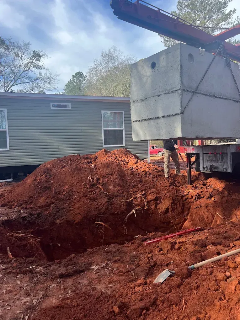 A crane lowers a concrete septic tank into a dug pit next to a mobile home. A worker is present.