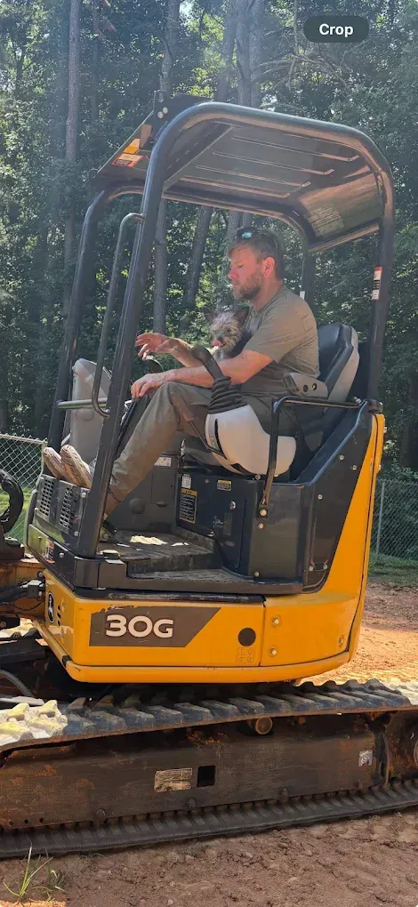 A person operating a yellow JCB 30G excavator in a wooded area.