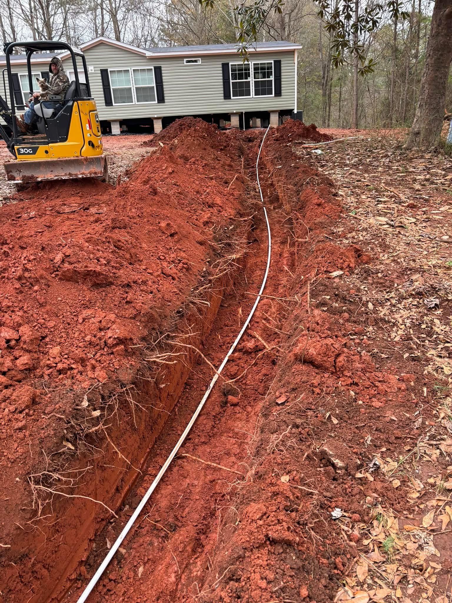 Trench dug in red soil, with white pipe laid, leading toward a house and small tractor.