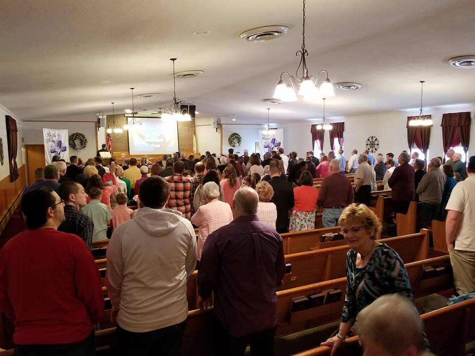 People standing in a church, facing forward. Light-colored walls and pews, projector screen in the distance.