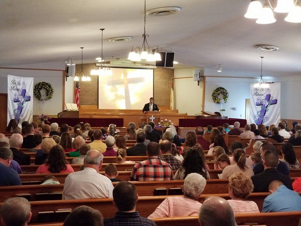 Church service: A pastor preaching from a pulpit, large congregation seated in pews. Cross projected on screen.