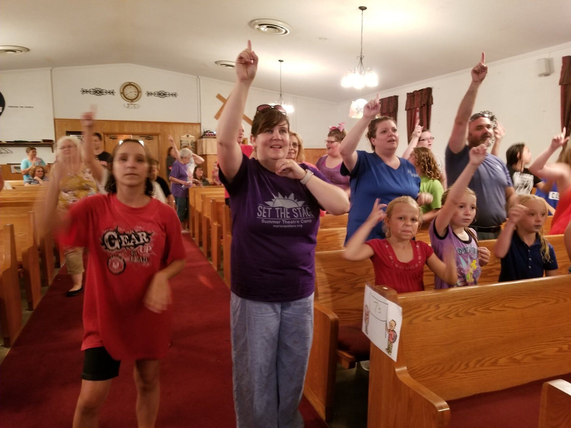 People in a church raise hands, singing. Interior of church with pews, red carpet, and a cross.