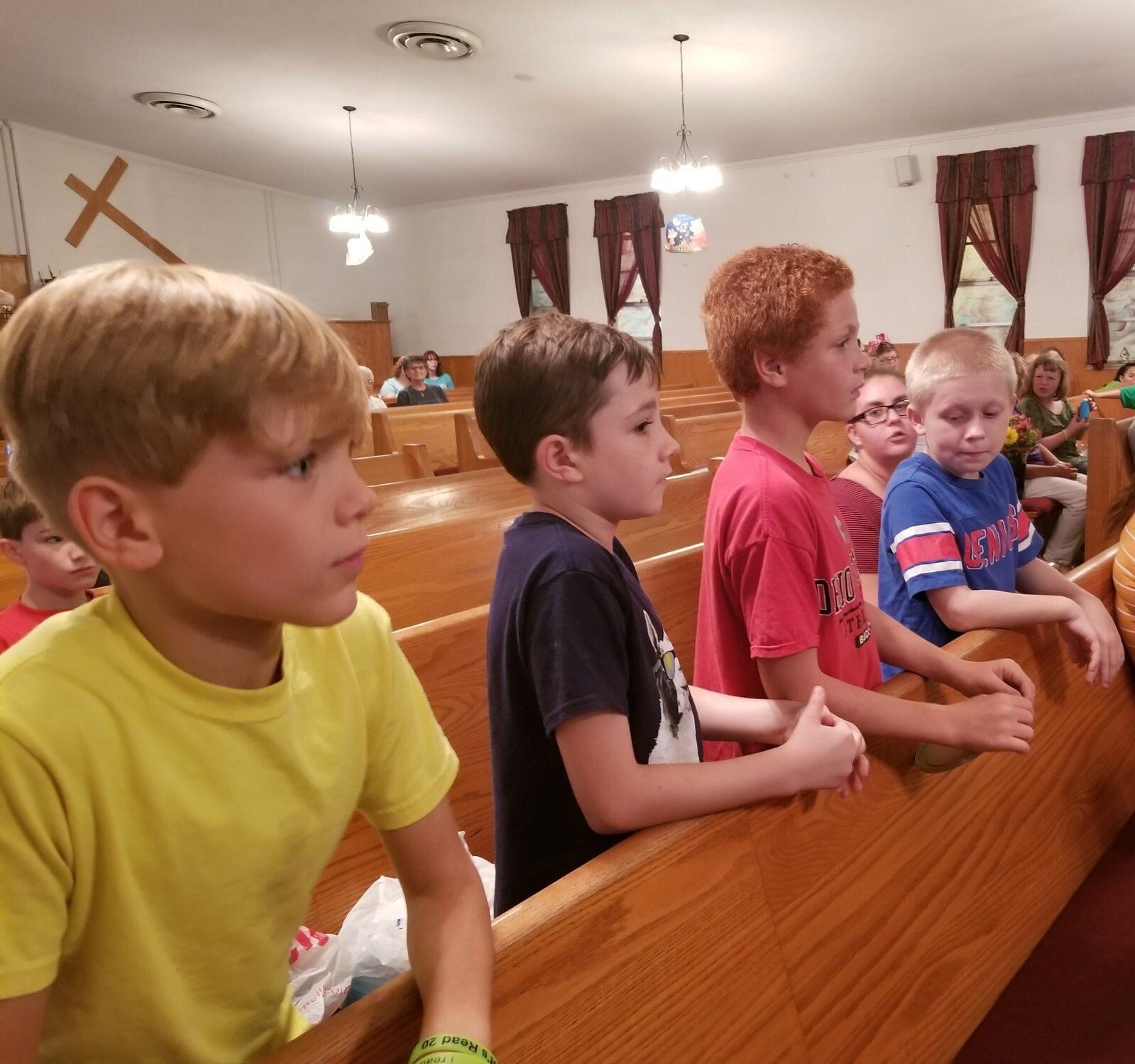Children in a church stand at pews, looking toward the front. Yellow, red, and blue shirts. Wooden interior, cross visible.