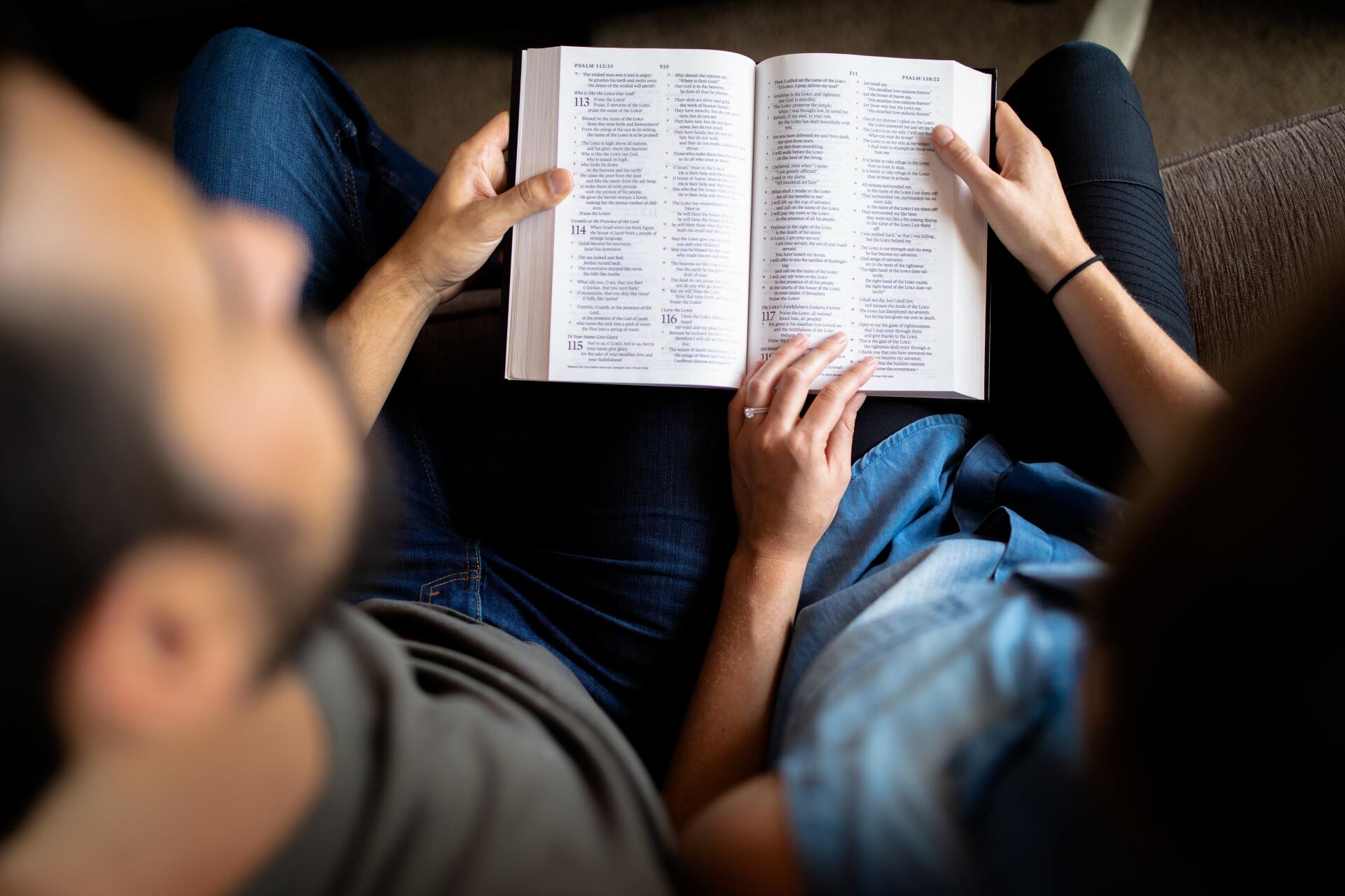 Couple reading a book together; seated, close-up. Focus on open book with text, hands pointing to words.