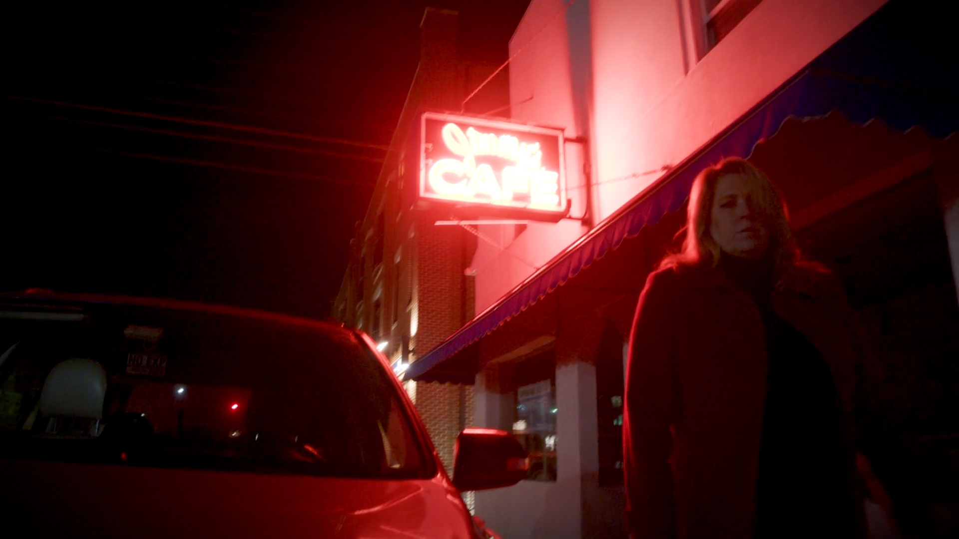 A woman is standing in front of a red car in front of a neon sign.