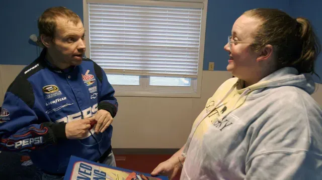 a man in a race car jacket is talking to a woman in a room .