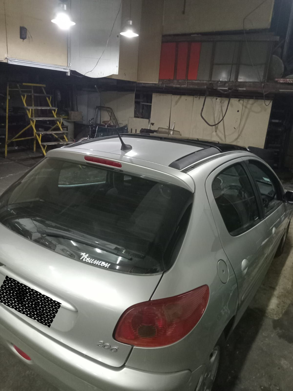 Un Peugeot 206 plateado estacionado en el interior de un taller o garaje, visto desde la parte trasera del lado del pasajero.