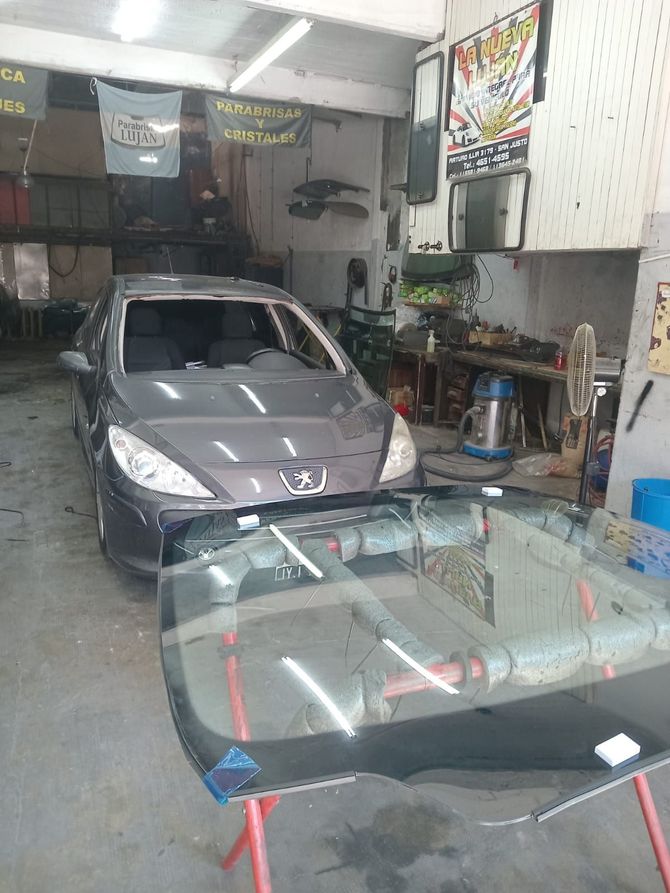 Un Peugeot gris oscuro aparcado en un taller mecánico, con un parabrisas de repuesto sobre un soporte en primer plano.