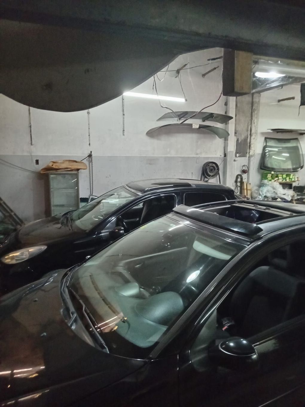 Dos coches oscuros aparcados dentro de un garaje, con piezas de automóviles colgando de la pared al fondo.