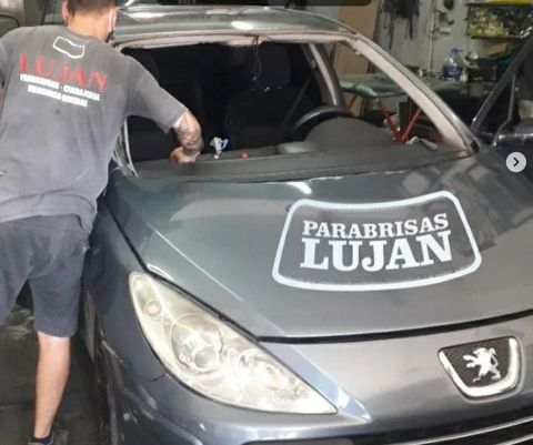 Un técnico con camisa gris trabaja en el marco del parabrisas de un coche Peugeot gris en el taller de Parabrisas Luján.