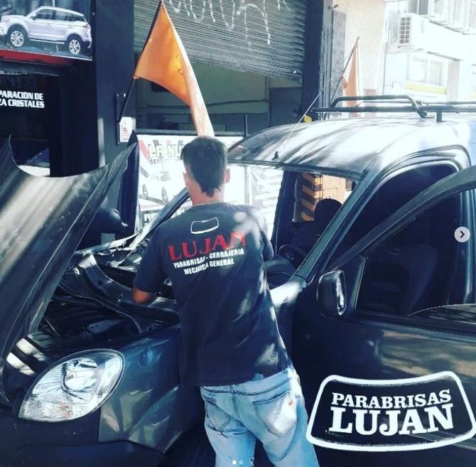 Un técnico con uniforme de marca trabaja en el parabrisas y el capó abierto de un coche en Parabrisas Luján.