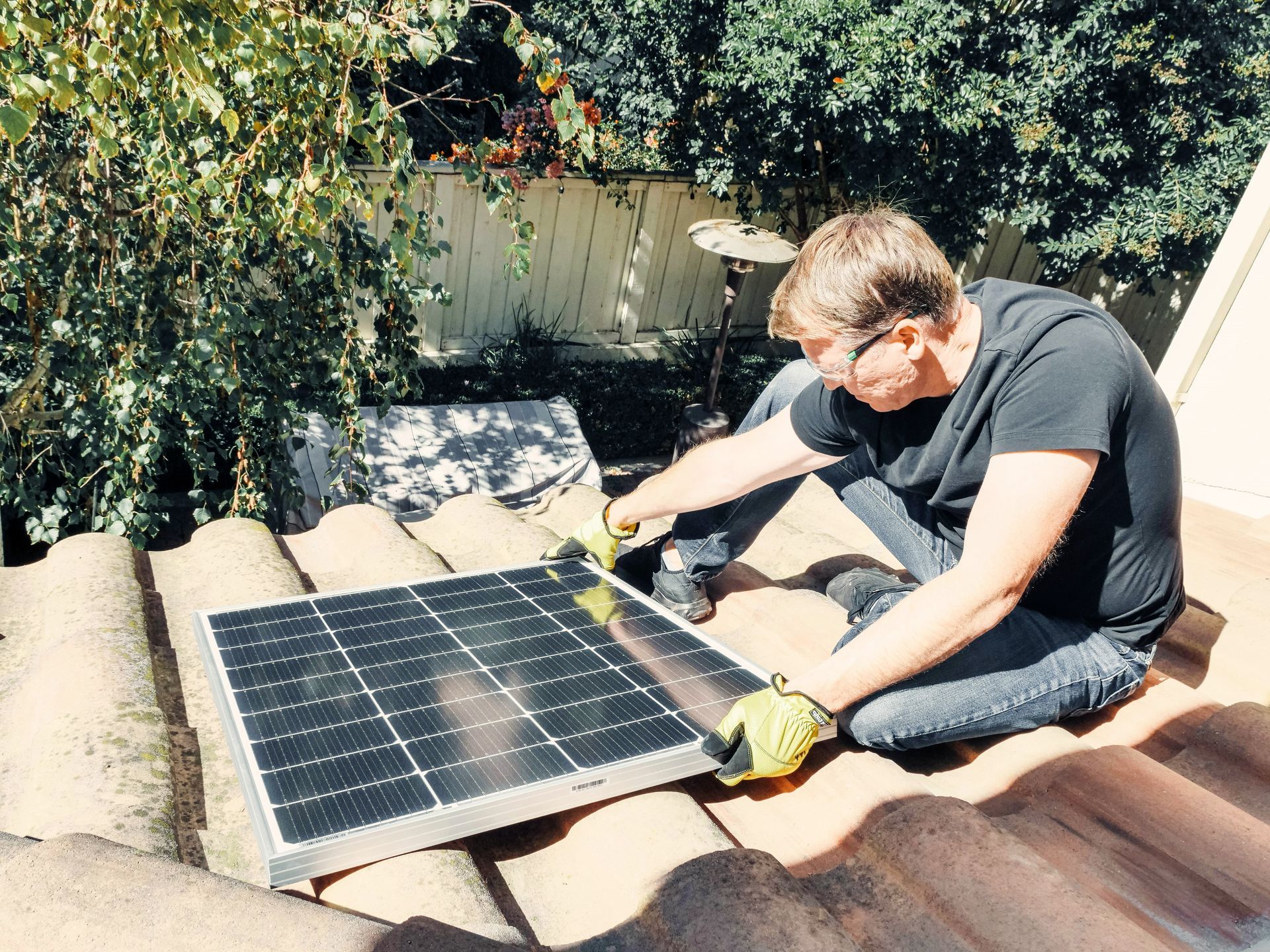 A man in a black shirt sits on a roof, holding a solar panel