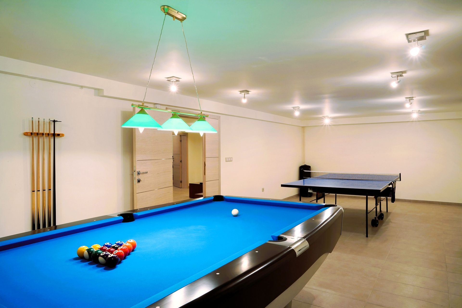 A game room with a blue billiard table, cue sticks, and a table tennis setup under bright lighting