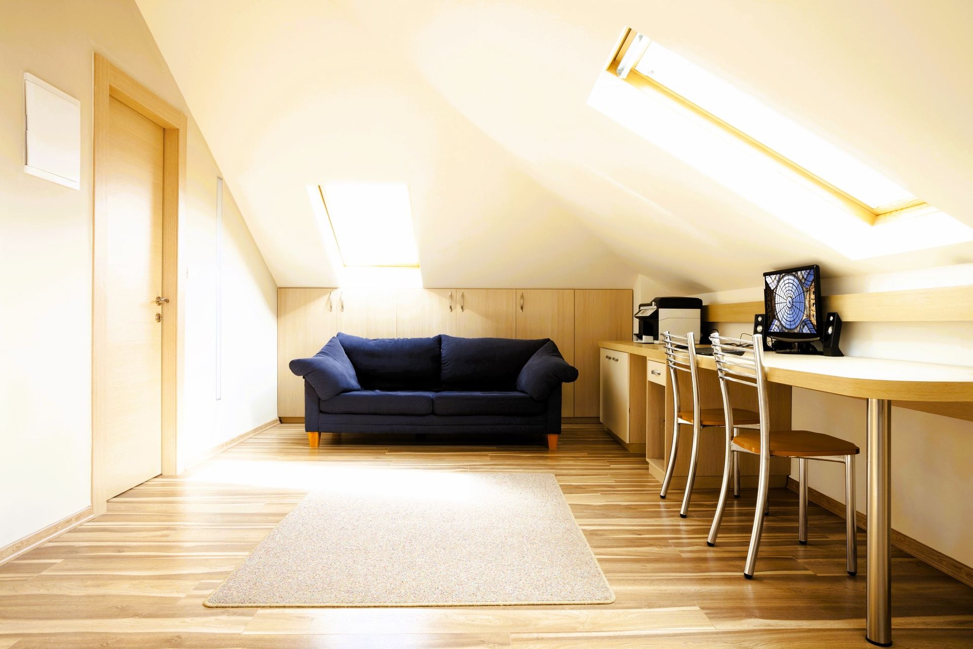 Cozy attic room with skylights, blue sofa, desk with computer, and beige carpet.