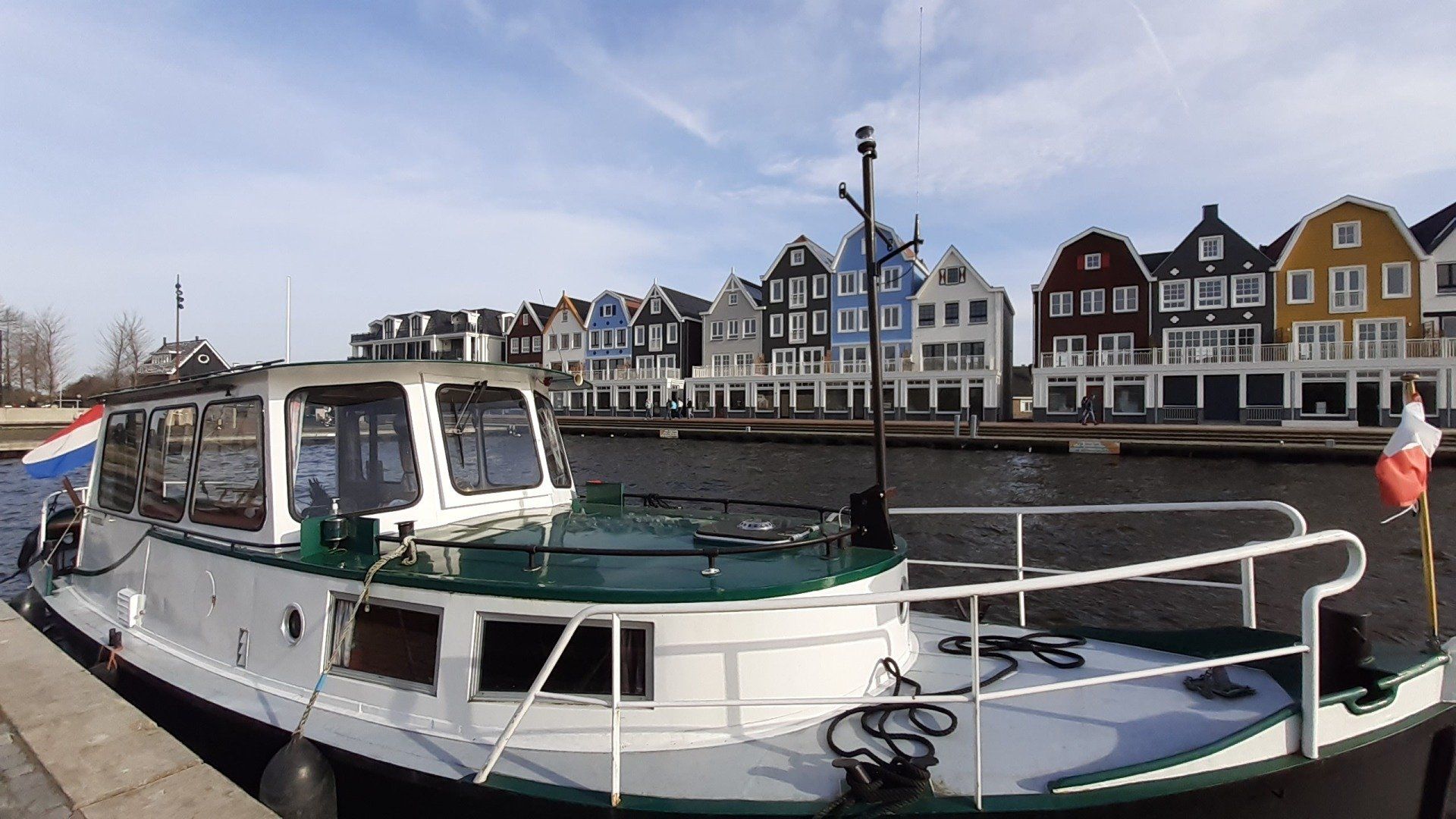 Een witte boot met een groen dak ligt aangemeerd in een kanaal voor kleurrijke traditionele gebouwen onder een blauwe hemel.