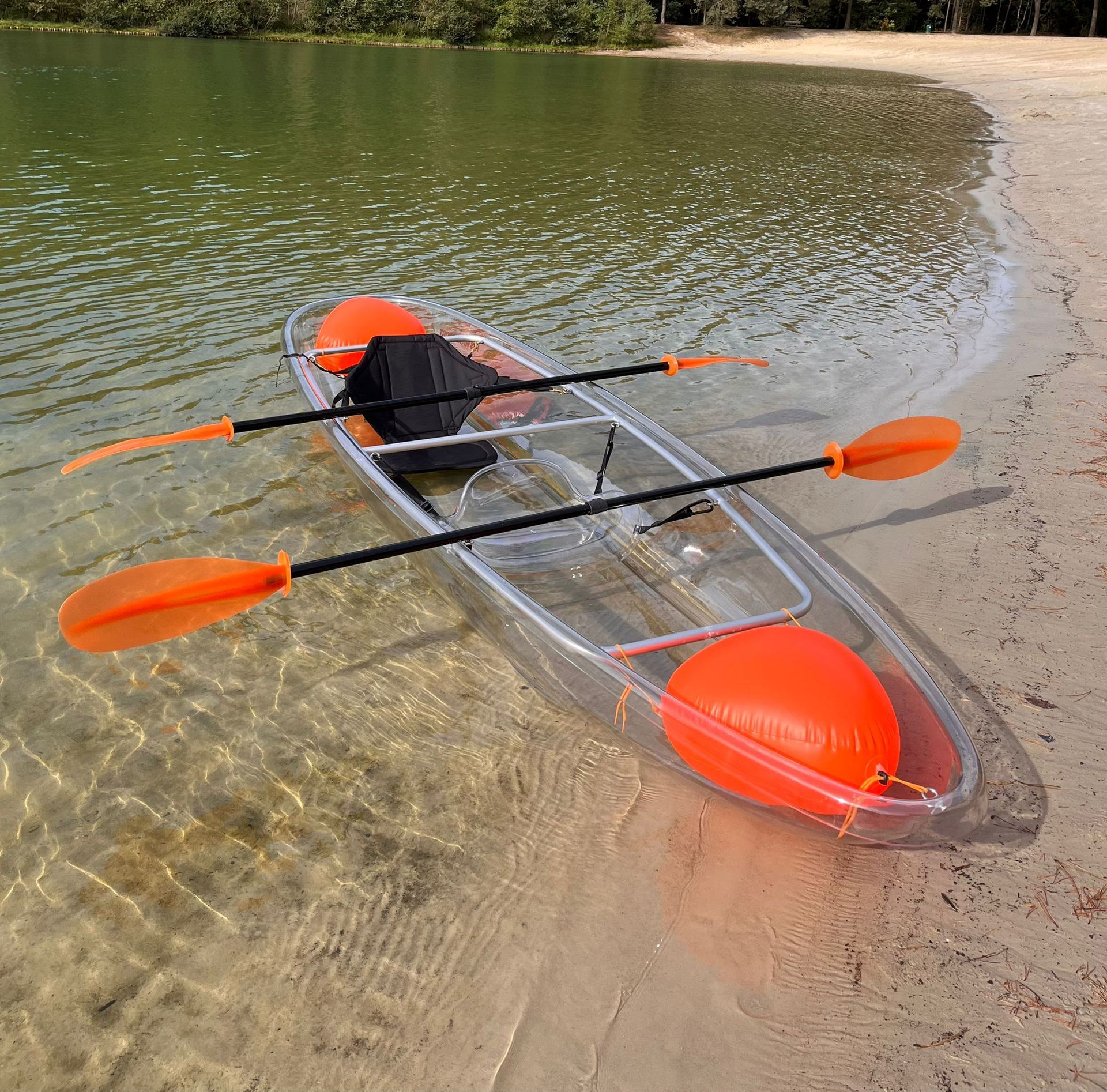 Een transparante kajak met oranje accenten en peddels op een zandstrand.