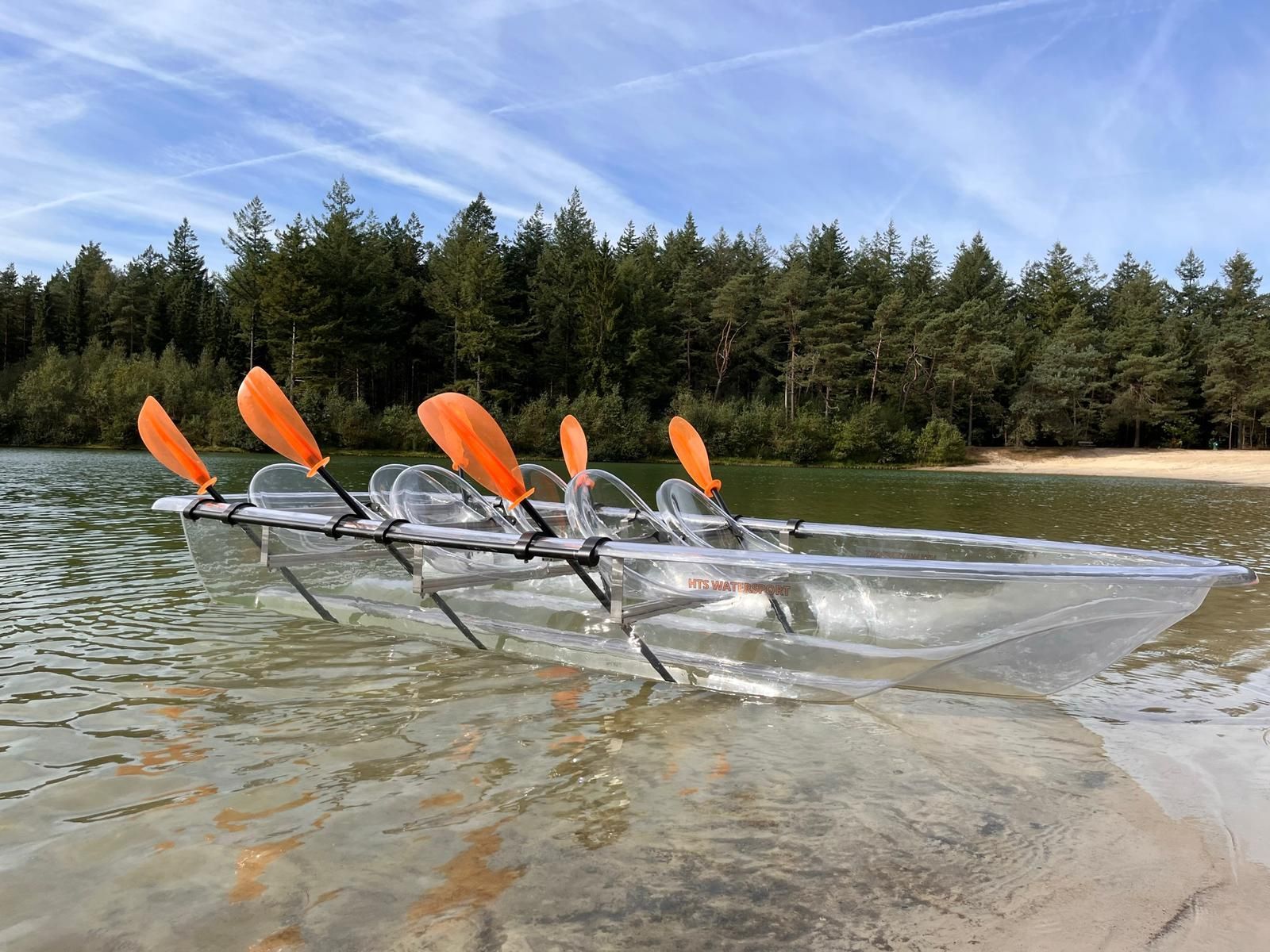 Een transparante kajak met oranje peddels drijft op het water, vlakbij een zandstrand en bomen.