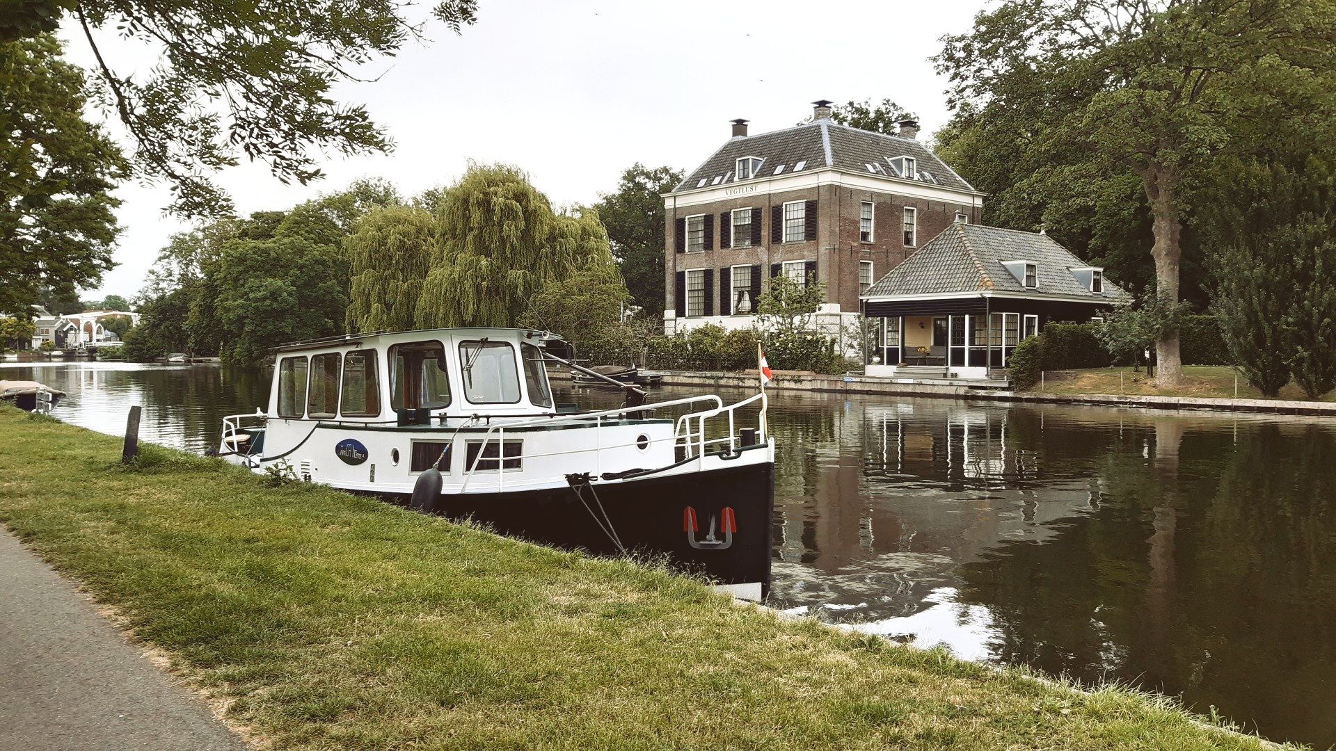 Een wit-zwarte boot ligt aangemeerd in een kanaal naast een klassiek bakstenen huis, omgeven door bomen.