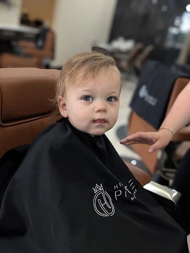 A toddler with blonde hair sits in a barber chair wearing a black cape. They look directly at the camera.