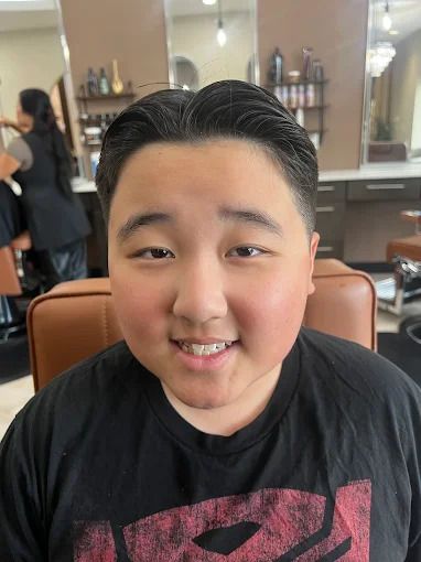Young person with short, slicked-back hair smiles at the camera in a salon setting.