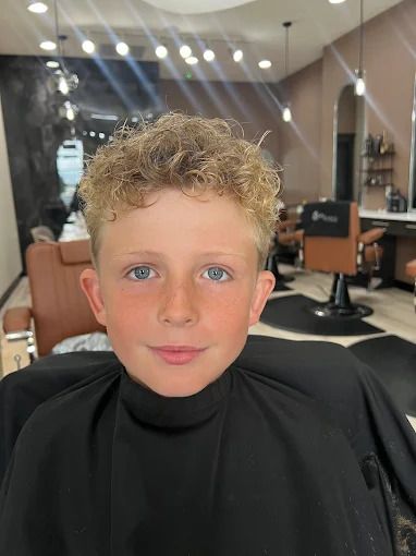 Boy with curly blond hair smiles at the camera in a barber shop after a haircut.
