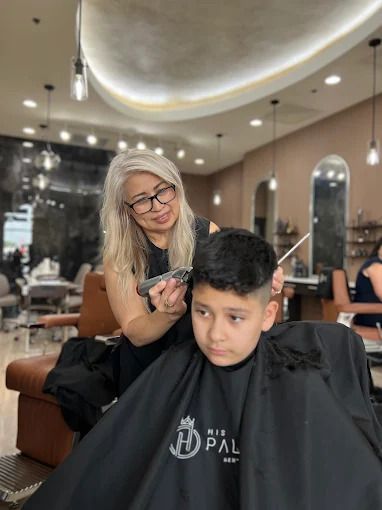 Hairdresser cutting a boy's hair with clippers in a salon. The boy is wearing a black cape, looking forward.