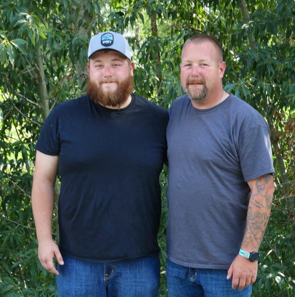 Two men standing side-by-side, smiling. One with a beard, a hat, and a black t-shirt. The other has short hair and a gray t-shirt.