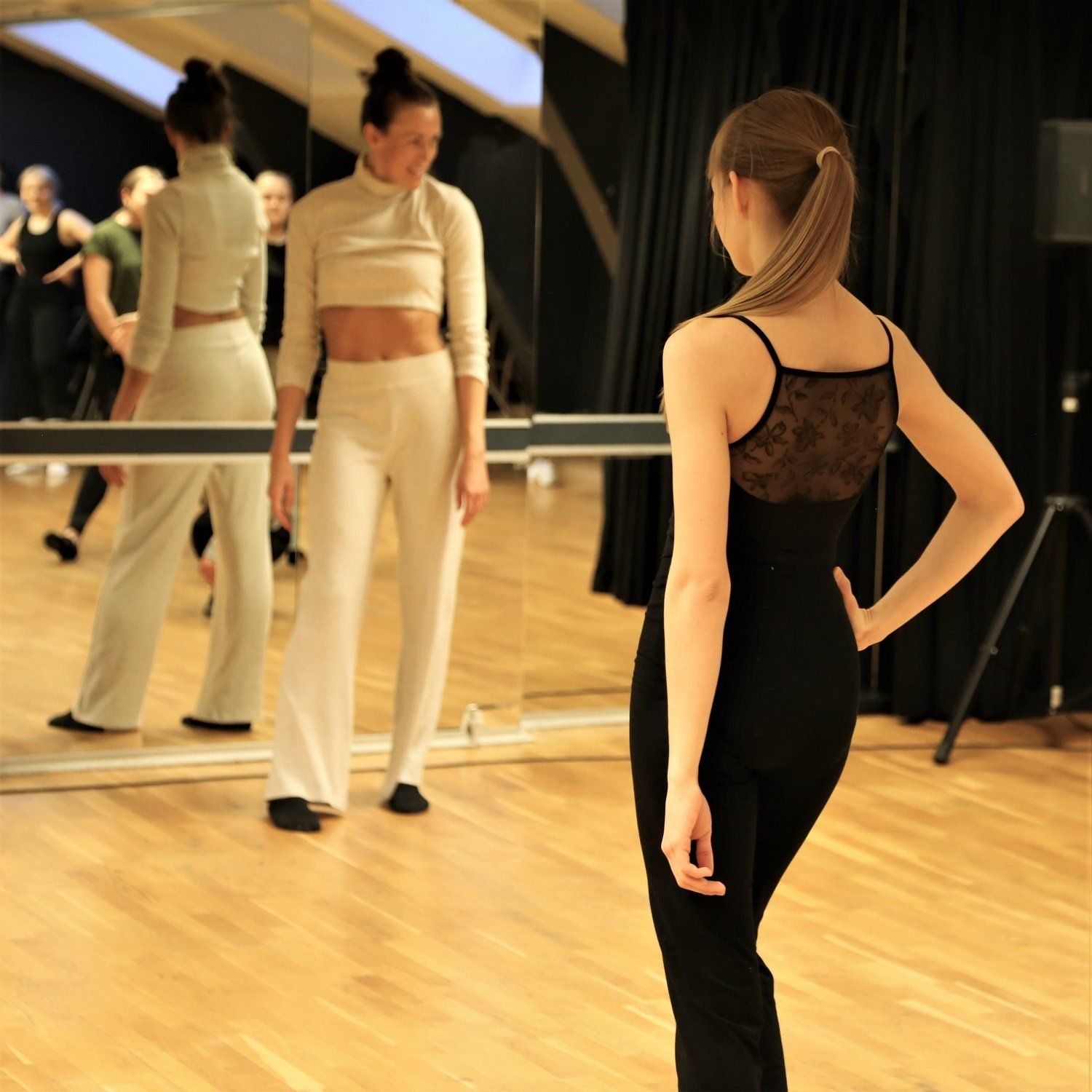 en kvinne i svart ballettdrakt står foran et speil i en dansesal