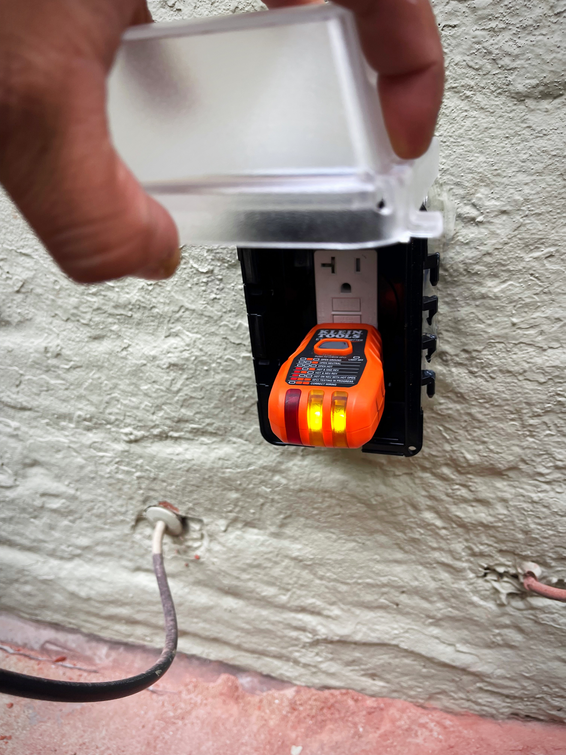 Hand opens a weatherproof cover over an outdoor electrical outlet, testing it with an orange circuit tester.