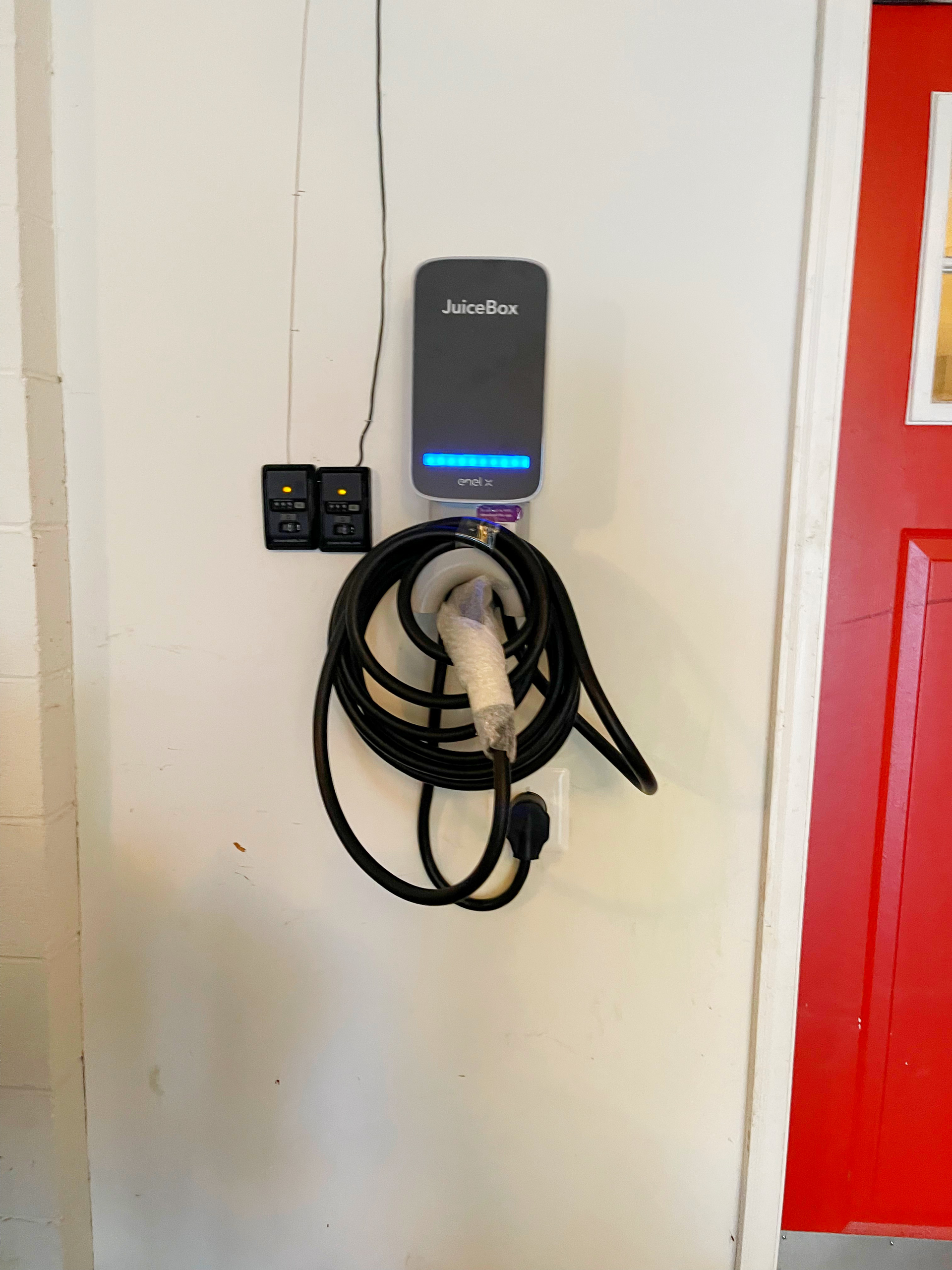 Electric vehicle charger mounted on a white wall next to a red door. Black cable coiled below.