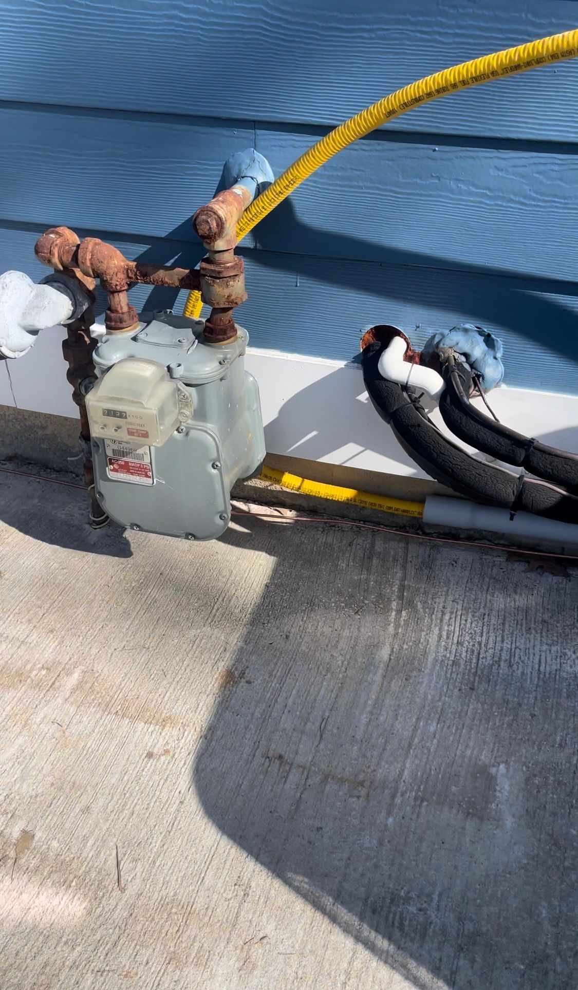 Gas meter on a blue house exterior with attached yellow hose and insulated black pipes on concrete.
