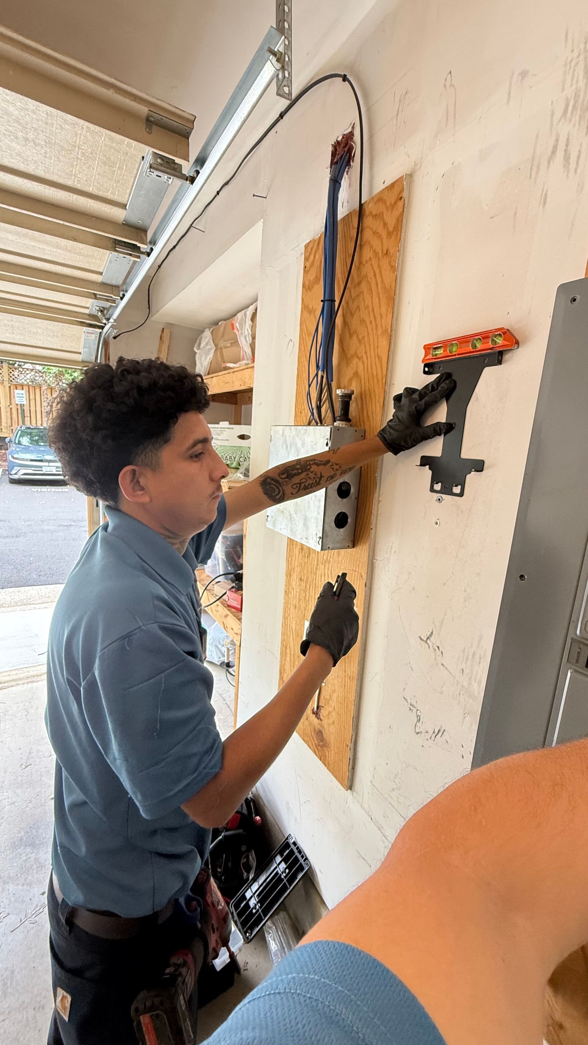 An electrician in blue shirt and gloves works on electrical wiring in a garage, using a tool.