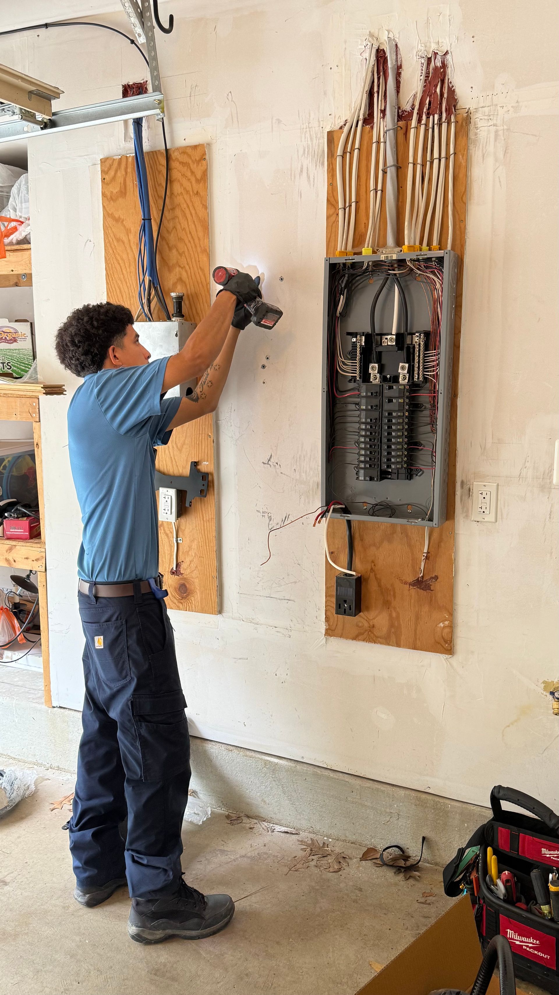 Electrician installing electrical panel in garage. Wears work clothes, using power tool.