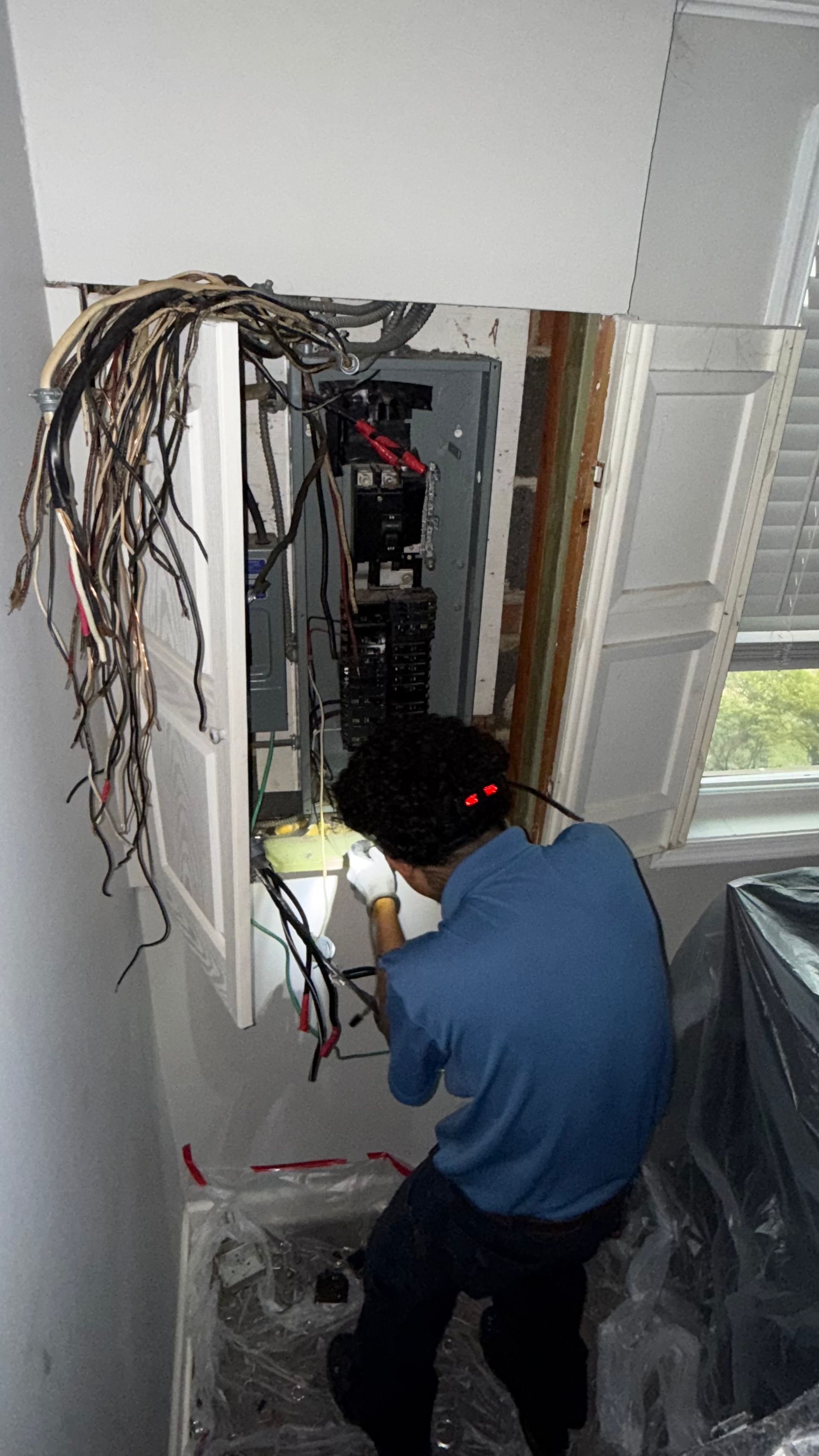 Electrician working on a circuit breaker panel, surrounded by wires. Inside a room with a window.