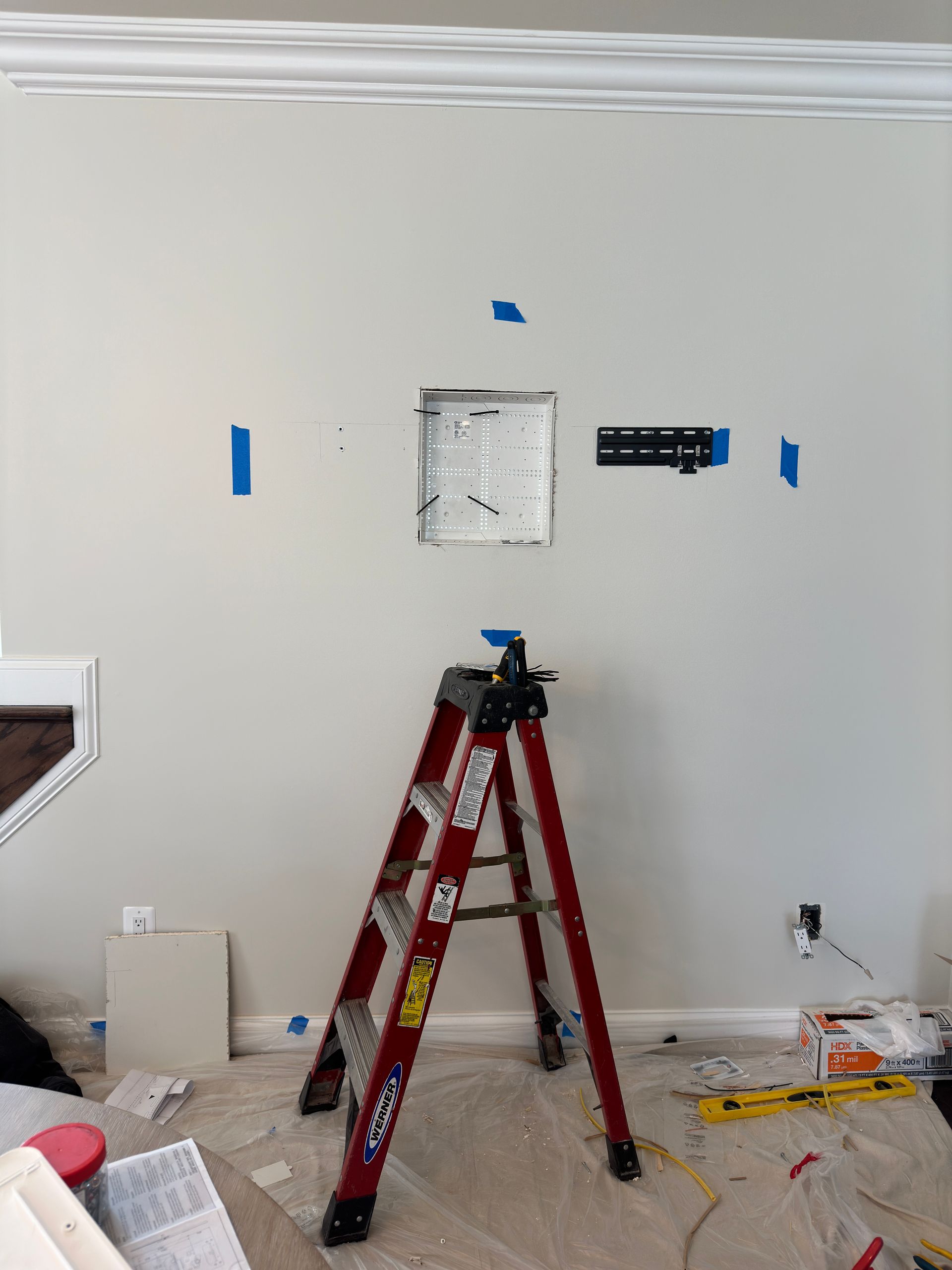 A ladder and electrical work in progress on a light-colored wall, with masking tape and a recessed box.